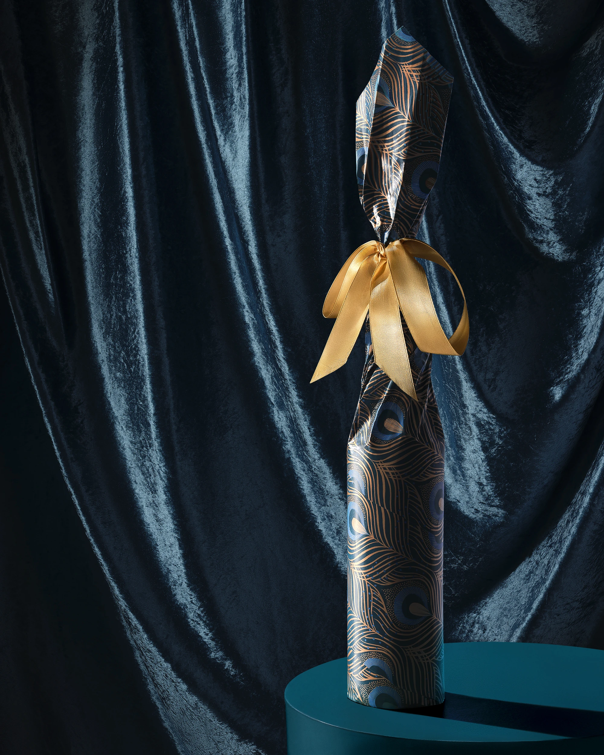 Commercial product photography of a bottle wrapped in peacock-patterned gift paper with a gold ribbon against dark teal velvet at a London studio.