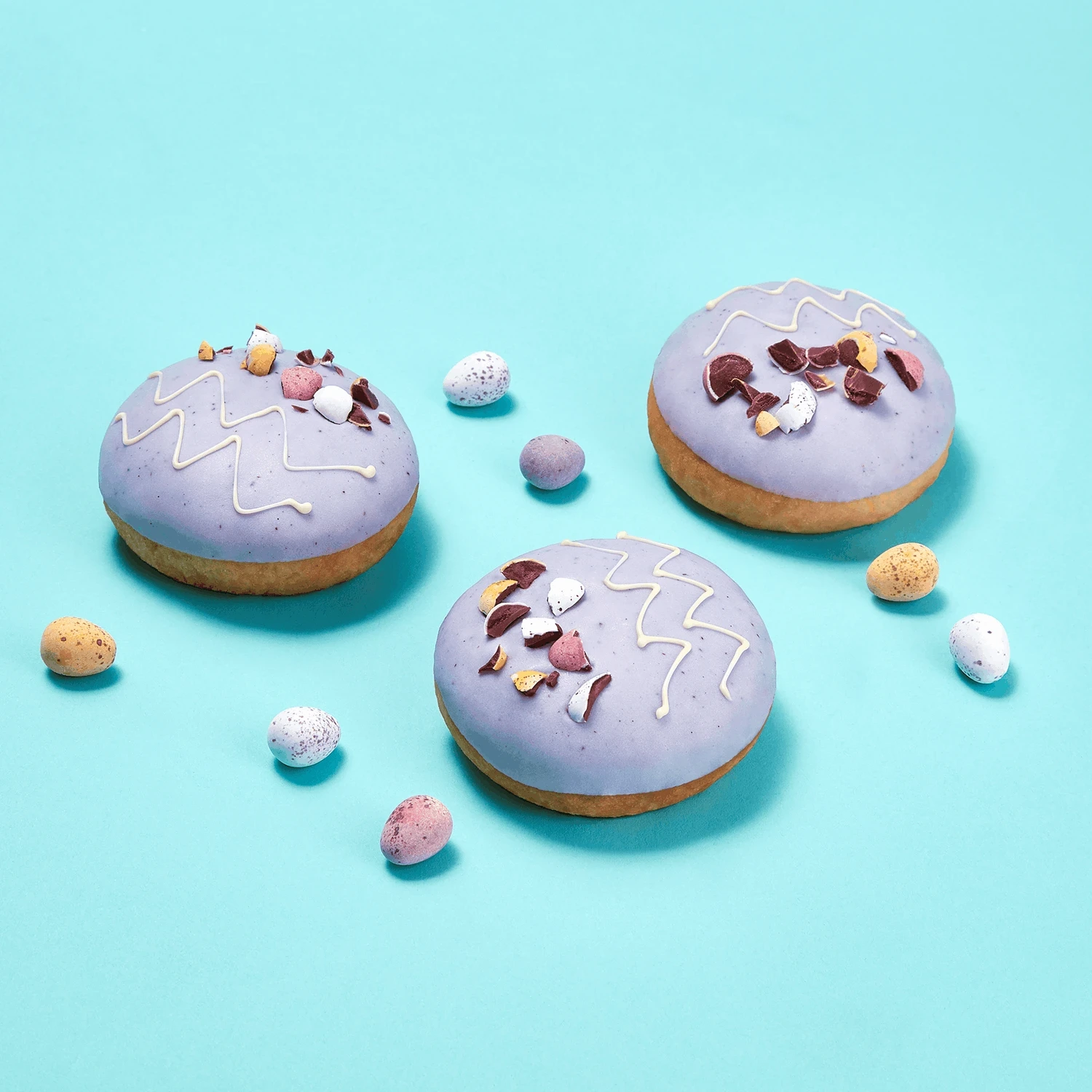 Creative food photography of purple-glazed Easter doughnuts decorated with mini chocolate eggs on a turquoise background.