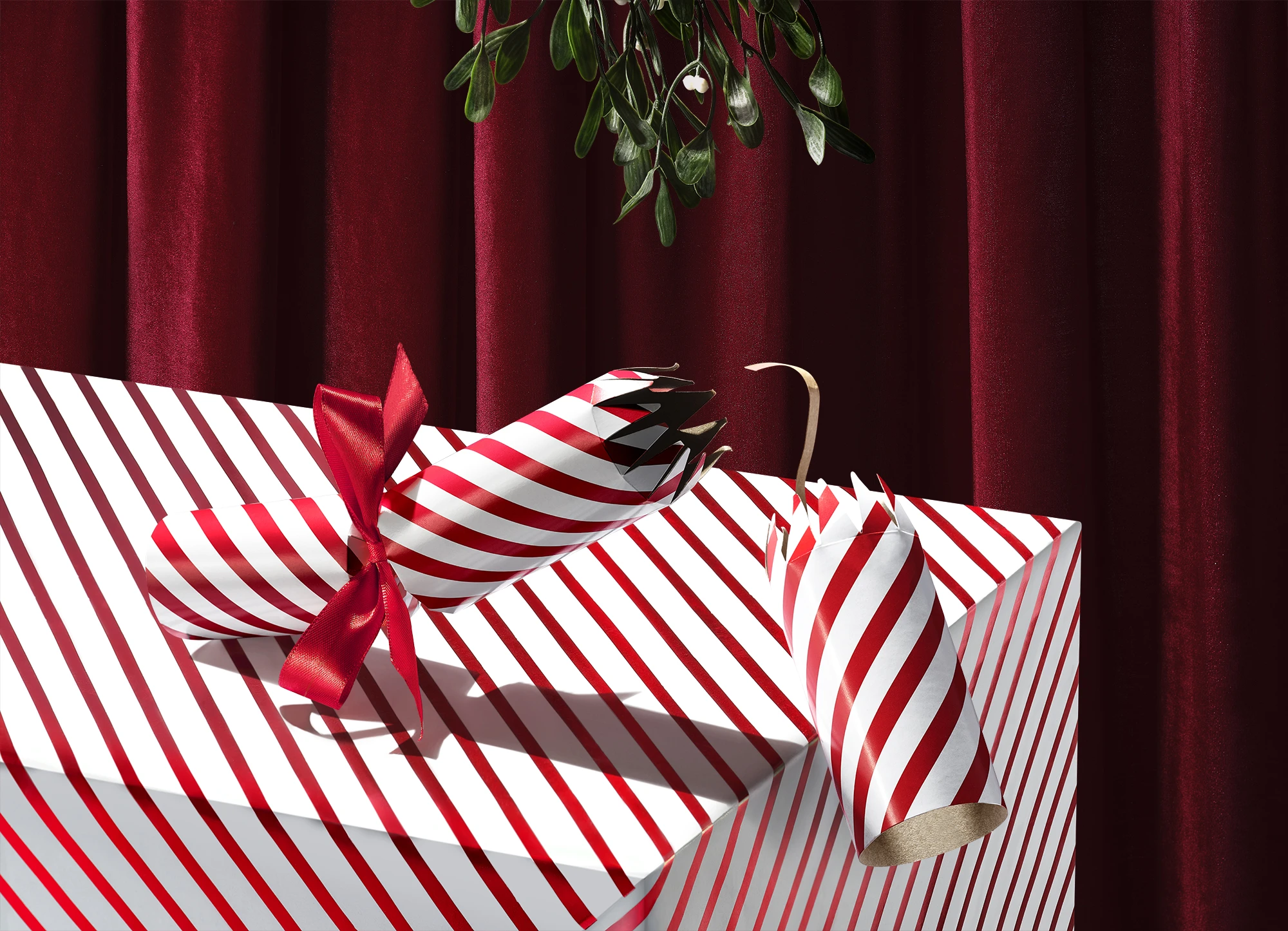 Commercial product photography of red-and-white striped Christmas crackers and gift boxes beneath mistletoe.