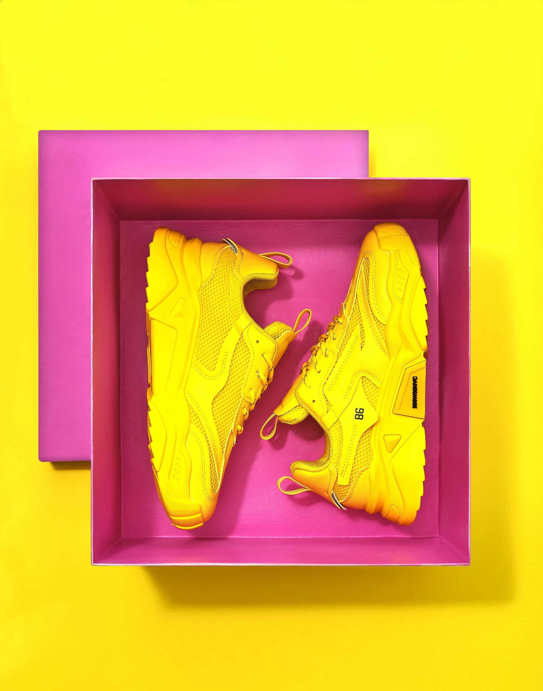 Fashion product photography of bright yellow chunky trainers arranged inside a pink display box.